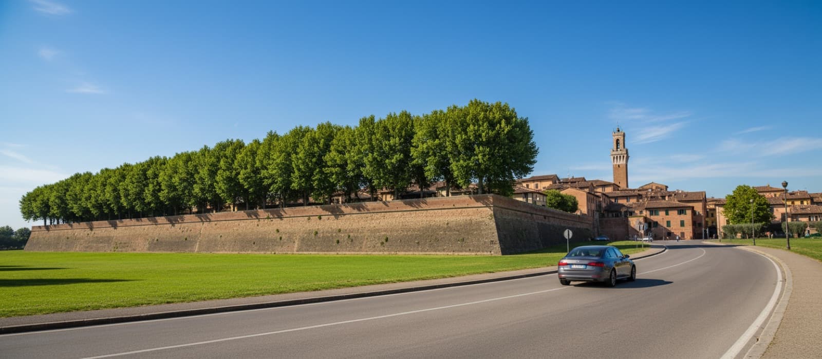 A car on the avenue beside tree-topped brick city ramparts, with rooftops and a tower behind